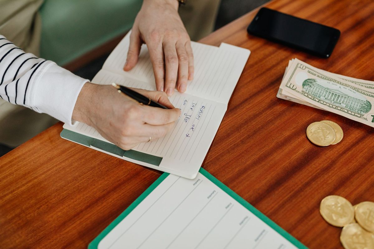 Hands writing a budget in a notebook next to cash and coins